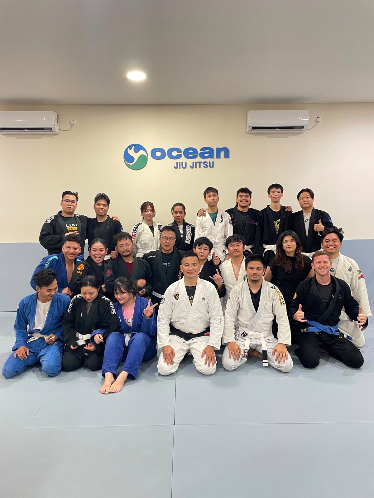 A group of students and instructors at a Brazilian Jiu-Jitsu academy, mixed belts, gathered for a class photo